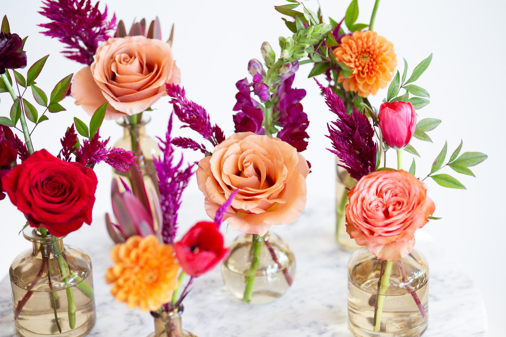 WildFlora's Gathered Garden Bud Vases in tan glass vessels on a marble table. They include orange, tangerine, red, pink, fuchsia, maroon, and gold flowers, complete with ranunculus, rose, dahlia, celosia, lisianthus, tulip, and snapdragon.