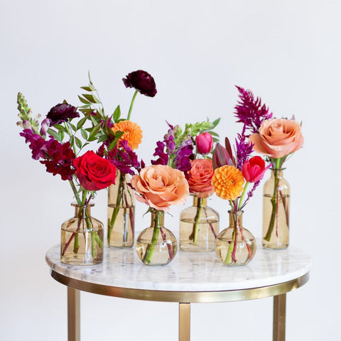 WildFlora's Gathered Garden Bud Vases in tan glass vessels on a marble table. They include orange, tangerine, red, pink, fuchsia, maroon, and gold flowers, complete with ranunculus, rose, dahlia, celosia, lisianthus, tulip, and snapdragon.