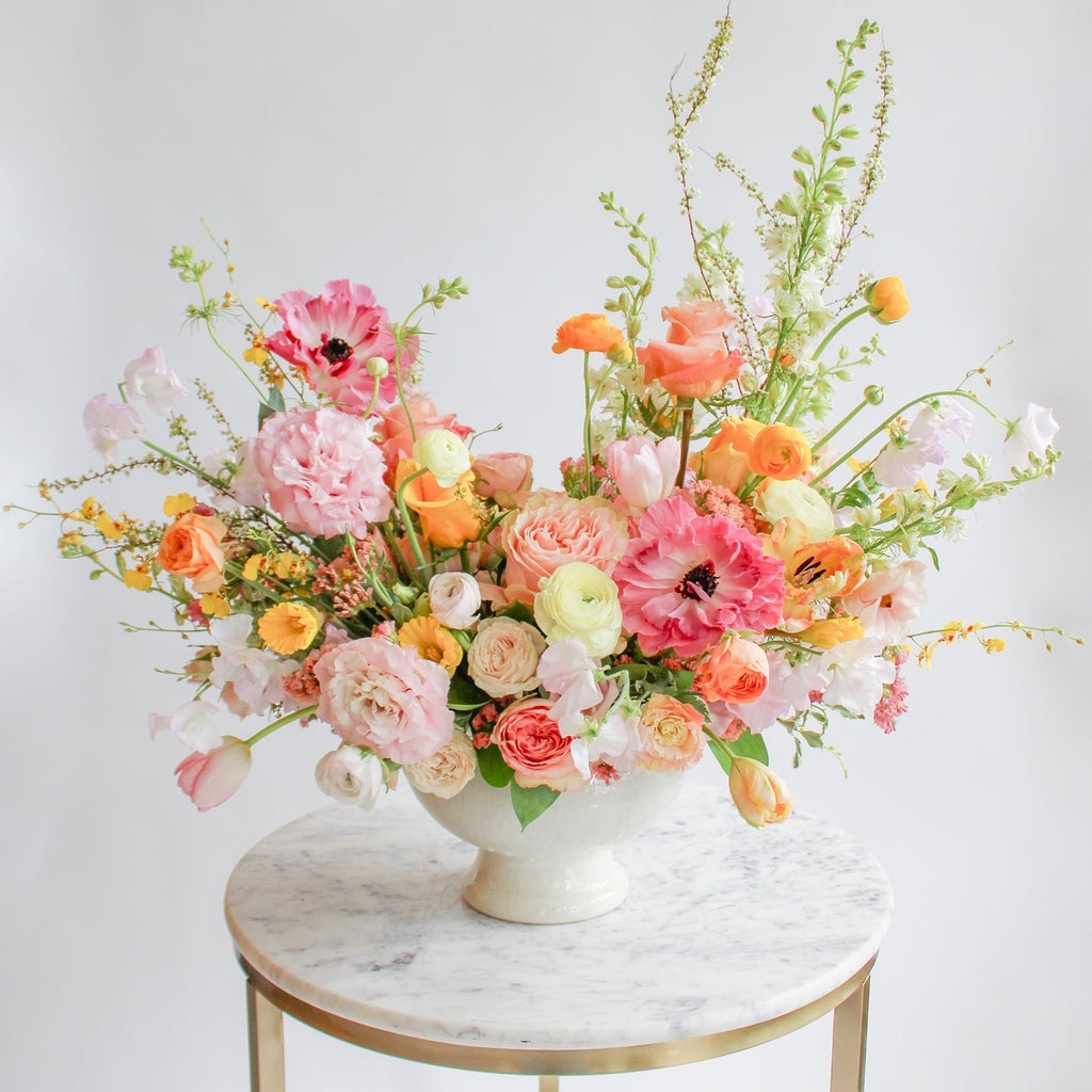 A flower arrangement in a glazed white compote vase in front of a white backdrop on a marble table. It's filled with pink, coral, yellow, peach, orange, and chartreuse-colored flowers, including rose, japanese ranunculus, daffodil, daisy, tulip, oncidium orchid, status, and delphinium. The vibes are very Bridgerton.