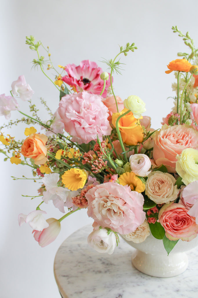 Close-up of a flower arrangement in a glazed white compote vase in front of a white backdrop on a marble table. It's filled with pink, coral, yellow, peach, orange, and chartreuse-colored flowers, including rose, japanese ranunculus, daffodil, daisy, tulip, oncidium orchid, status, and delphinium. The vibes are very Bridgerton.