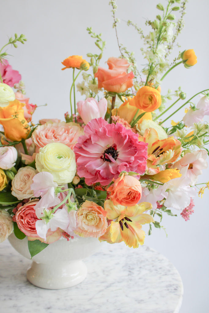 Close-up of a flower arrangement in a glazed white compote vase in front of a white backdrop on a marble table. It's filled with pink, coral, yellow, peach, orange, and chartreuse-colored flowers, including rose, japanese ranunculus, daffodil, daisy, tulip, oncidium orchid, status, and delphinium. The vibes are very Bridgerton.