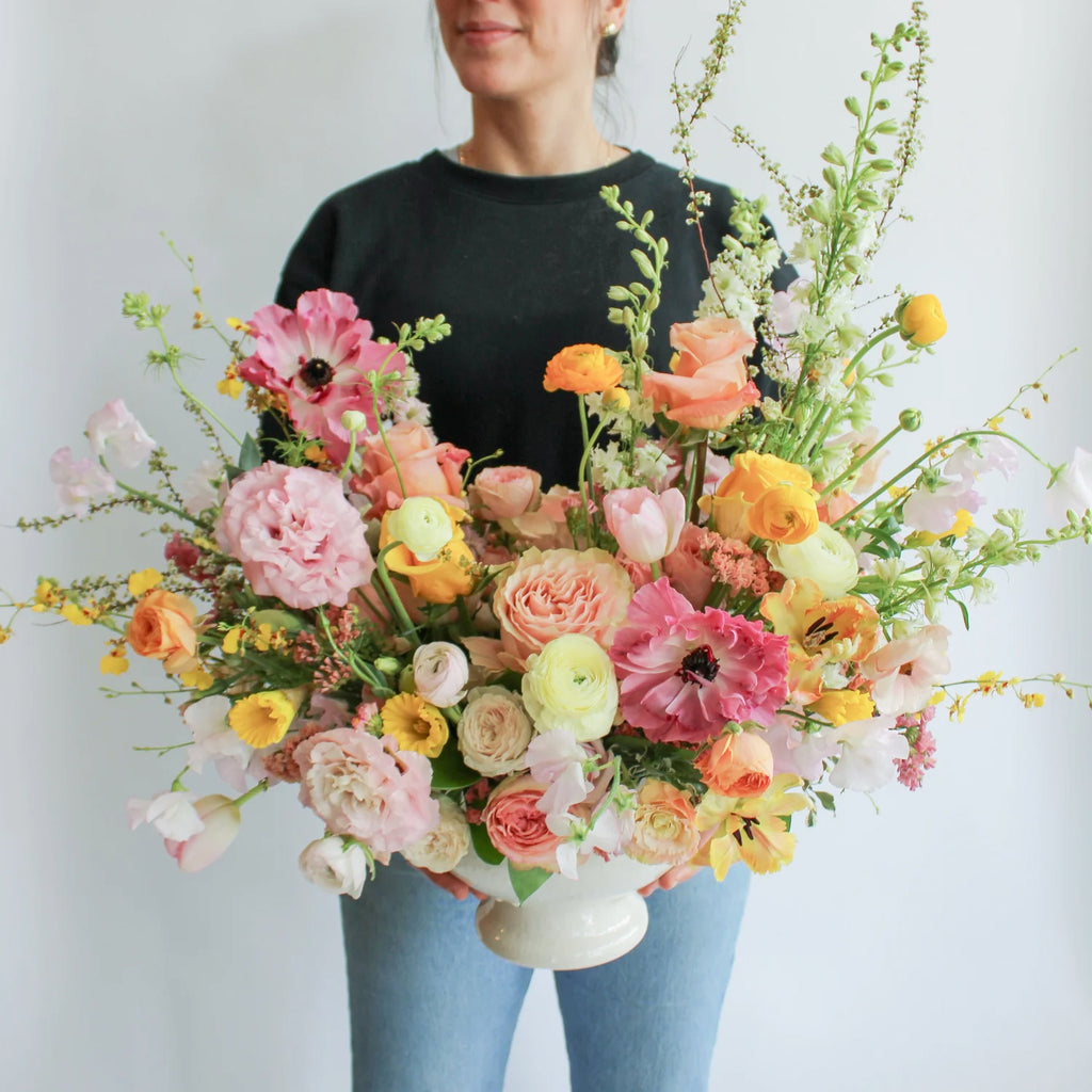 A woman holds a flower arrangement in a glazed white compote vase in front of a white backdrop. It's filled with pink, coral, yellow, peach, orange, and chartreuse-colored flowers, including rose, japanese ranunculus, daffodil, daisy, tulip, oncidium orchid, status, and delphinium. The vibes are very Bridgerton.