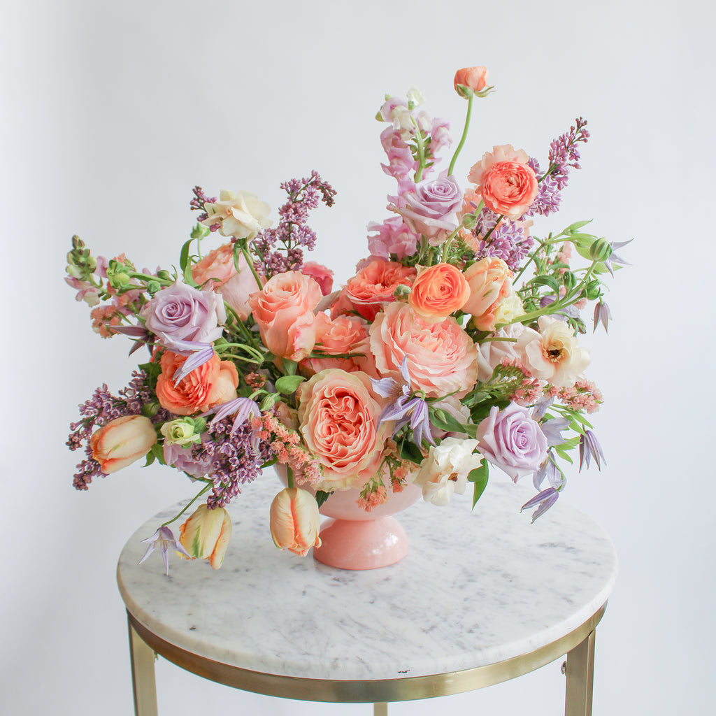 A flower arrangement in a pink vase on a marble table with peach, orange, purple, and lavender flowers, including rose, clematis, hyacinth, delphinium, anemone, tulip, and status. The backdrop is white.