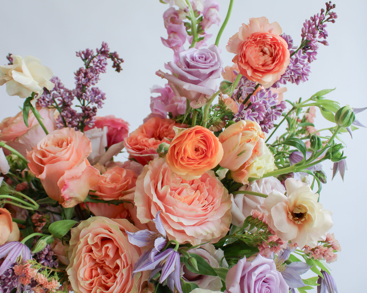 Close-up of a flower arrangement in a pink vase on a marble table with peach, orange, purple, and lavender flowers, including rose, clematis, hyacinth, delphinium, anemone, tulip, and status. The backdrop is white.