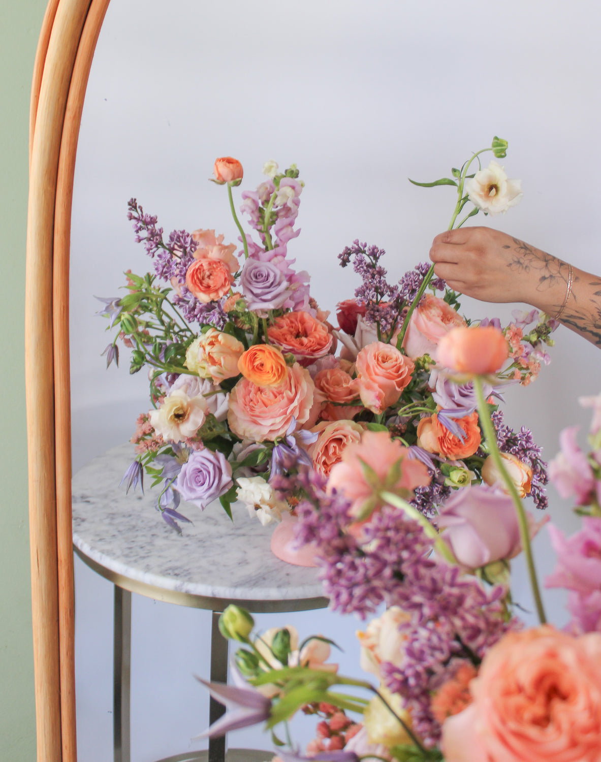 A reflection in a mirror of A flower arrangement in a pink vase on a marble table with peach, orange, purple, and lavender flowers, including rose, clematis, hyacinth, delphinium, anemone, tulip, and status. 