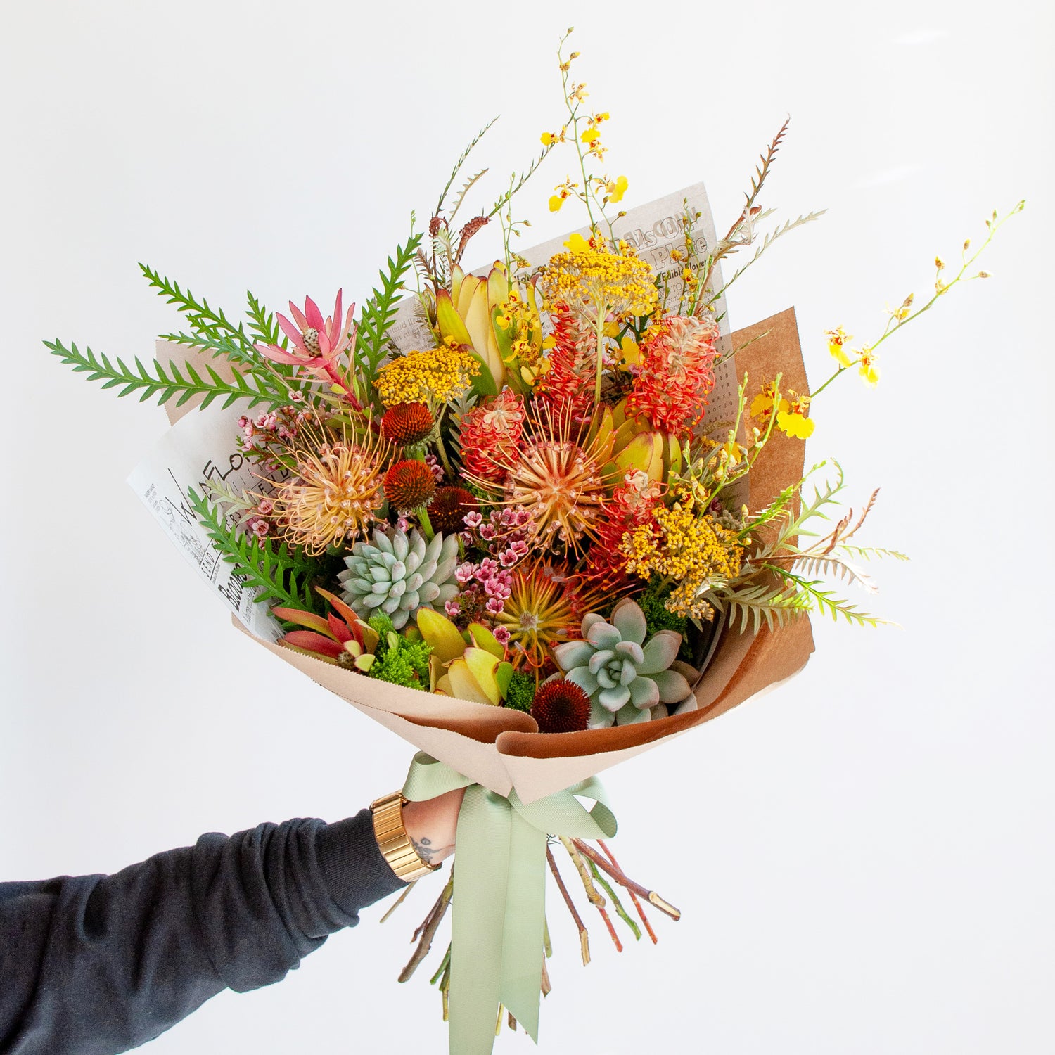 An arm holds a bouquet wrapped in floral newspaper and brown kraft paper in front of a white backdrop. It's filled with yellow, red, and orange flowers, including pincushion protea, grevillea, succulents, leucantha, oncidium orchid, and thistle. The bouquet has a green bow.