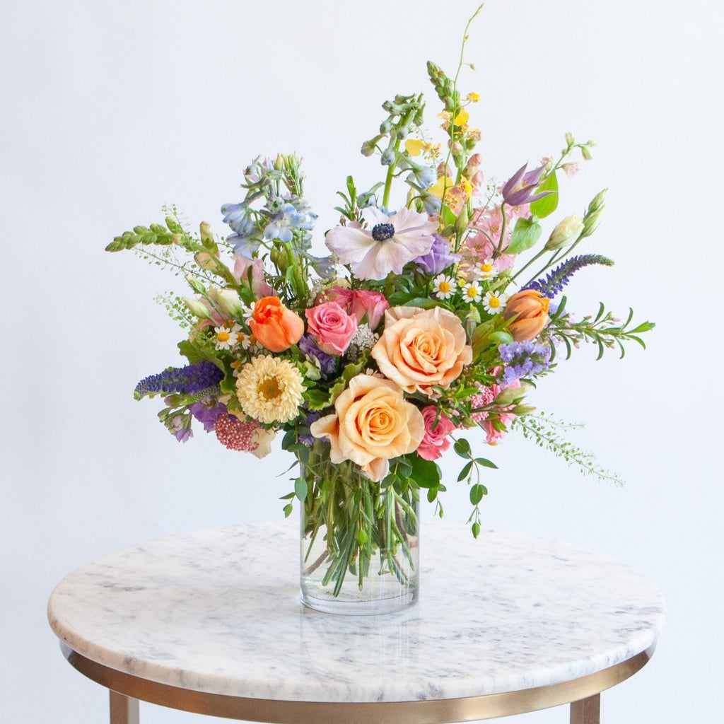 A glass vase with a petite flower arrangement sits on a marble table in front of a white backdrop. The flowers are many colors and include roses, tulips, snapdragon, veronica, anemone, clematis, orchids, and delphinium.