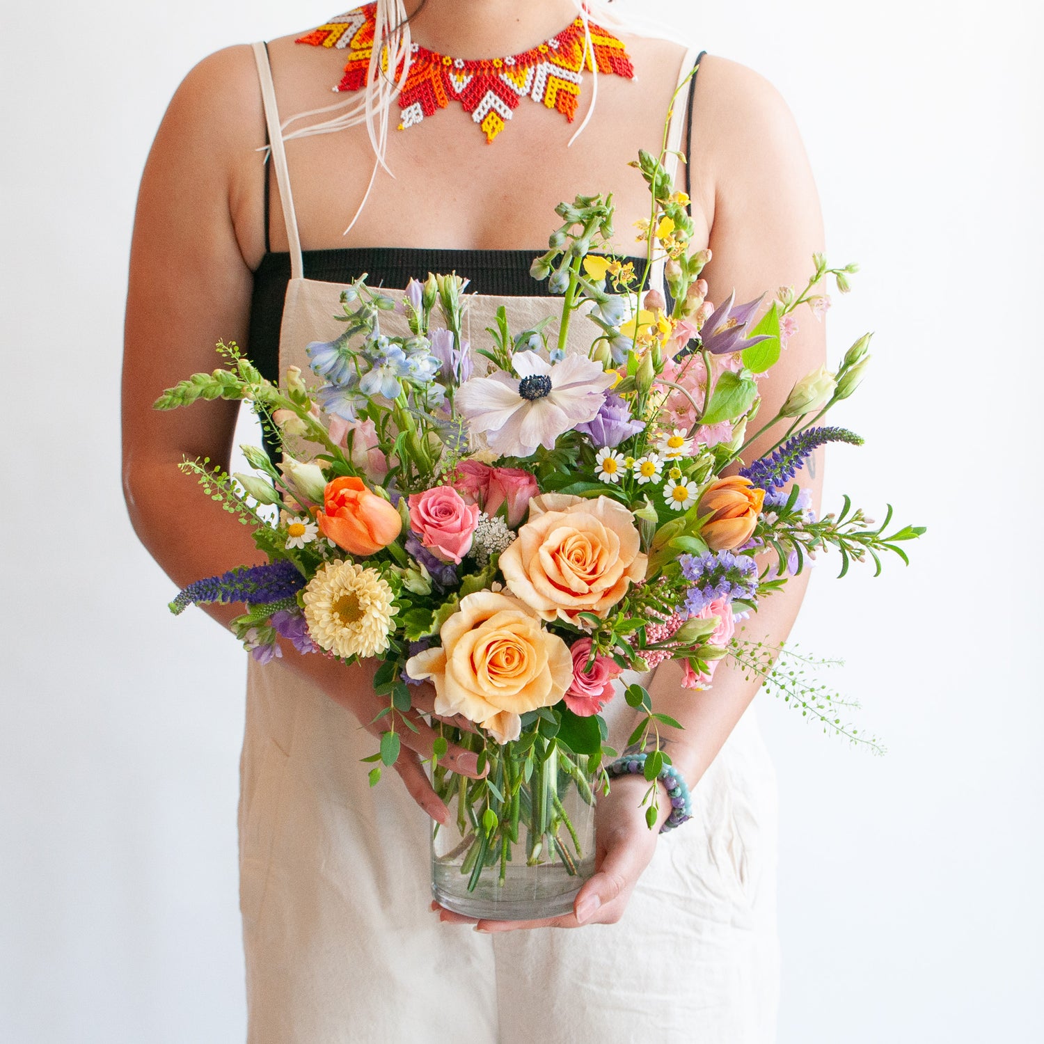A woman in cream overalls holds a glass vase with a petite flower arrangement in it.  The flowers are many colors and include roses, tulips, snapdragon, veronica, anemone, clematis, orchids, and delphinium.