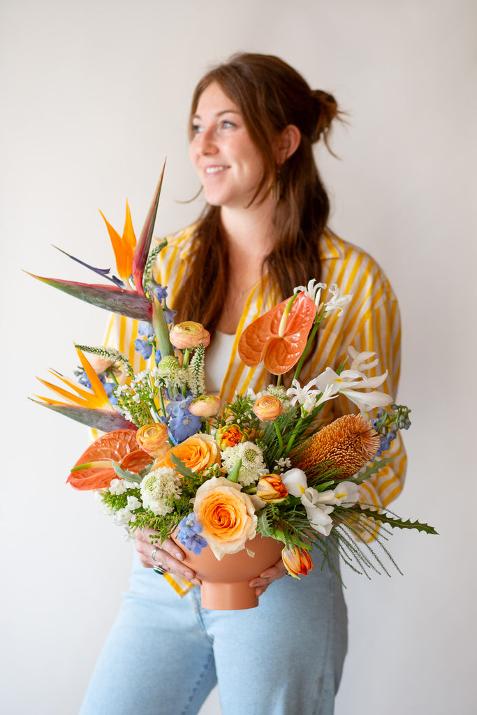 A woman holds a WildFlora's Tropic Paradise Arrangement in a terracotta compote vase in front of a white backdrop. It has blue, orange, peach, and white flowers, and includes rose, ranunculus, banksia protea, iris, delphinium, anthurium, scabiosa, bird of paradise, tulip, and allium.