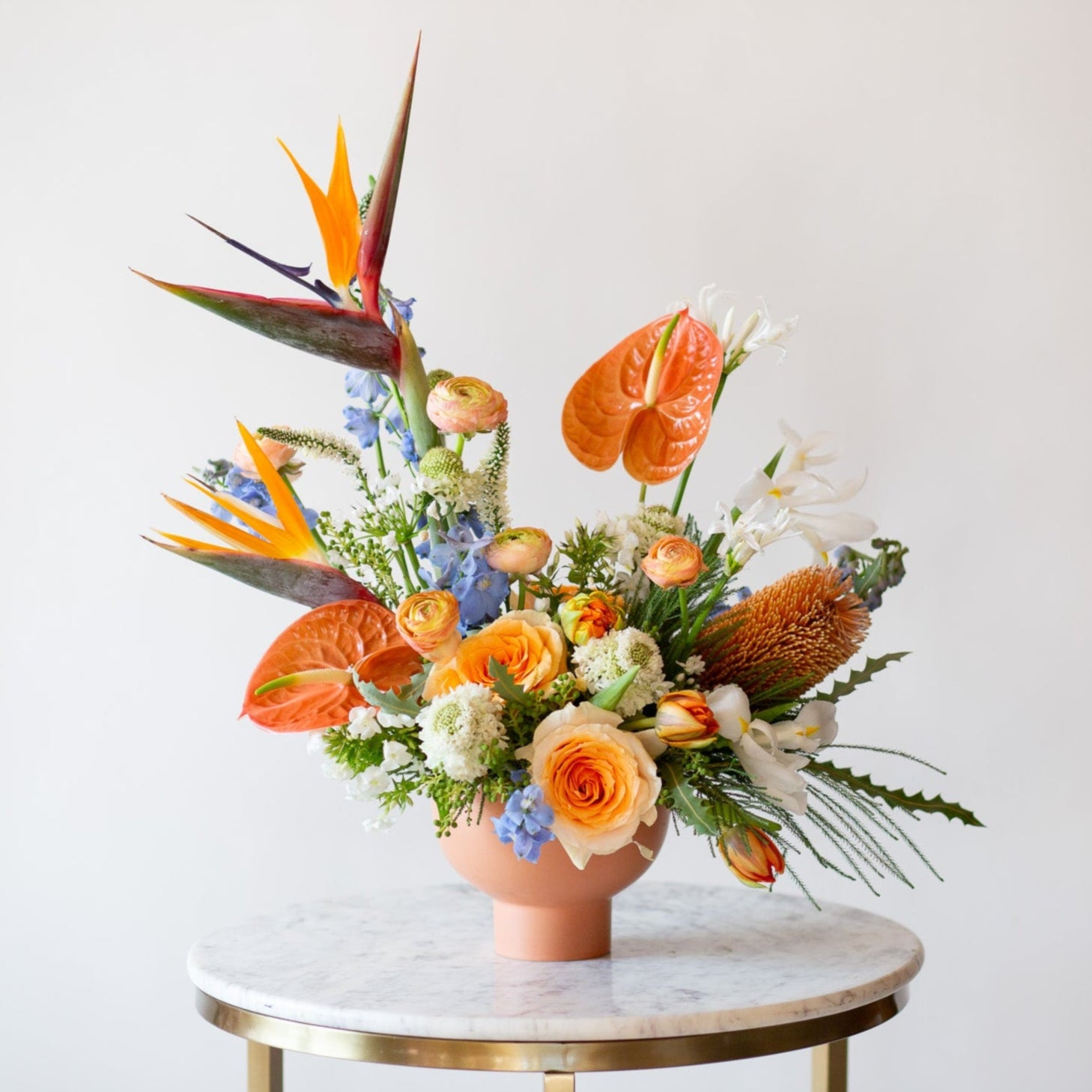 WildFlora's Tropic Paradise Arrangement in a terracotta compote vase on a small marble table in front of a white backdrop. It has blue, orange, peach, and white flowers, and includes rose, ranunculus, banksia protea, iris, delphinium, anthurium, scabiosa, bird of paradise, tulip, and allium.