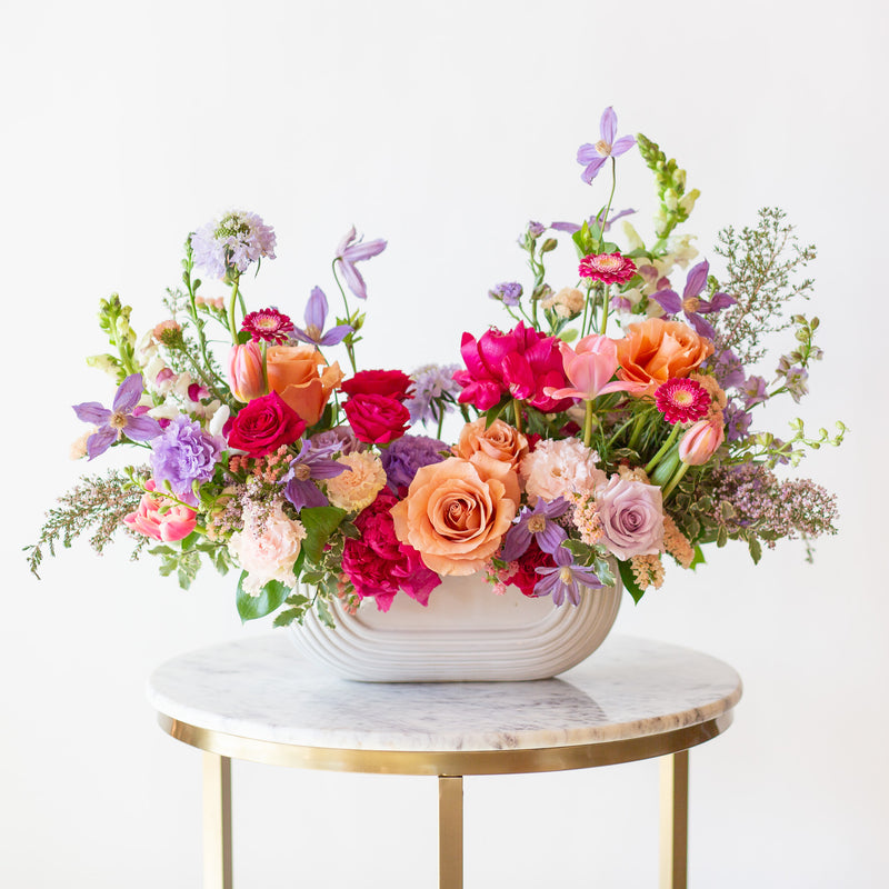 WildFlora's Wild Berry Garden Long & Low Arrangement in a white ceramic art deco long & low vase on a small marble table in front of a white backdrop. It has periwinkle, pink, magenta, orange, peach, purple and and white flowers, and includes rose, gerber daisy, lisianthus, snapdragon, clematis, scabiosa, peony, and tulips.