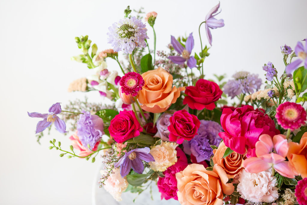 WildFlora's Wild Berry Garden Long & Low Arrangement in a white ceramic art deco long & low vase on a small marble table in front of a white backdrop. It has periwinkle, pink, magenta, orange, peach, purple and and white flowers, and includes rose, gerber daisy, lisianthus, snapdragon, clematis, scabiosa, peony, and tulips.