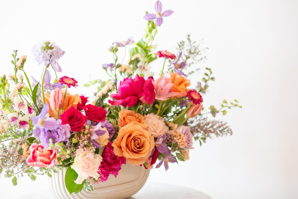 WildFlora's Wild Berry Garden  Long & Low Arrangement in a white ceramic art deco long & low vase on a small marble table in front of a white backdrop. It has periwinkle, pink, magenta, orange, peach, purple and and white flowers, and includes rose, gerber daisy, lisianthus, snapdragon, clematis, scabiosa, peony, and tulips.