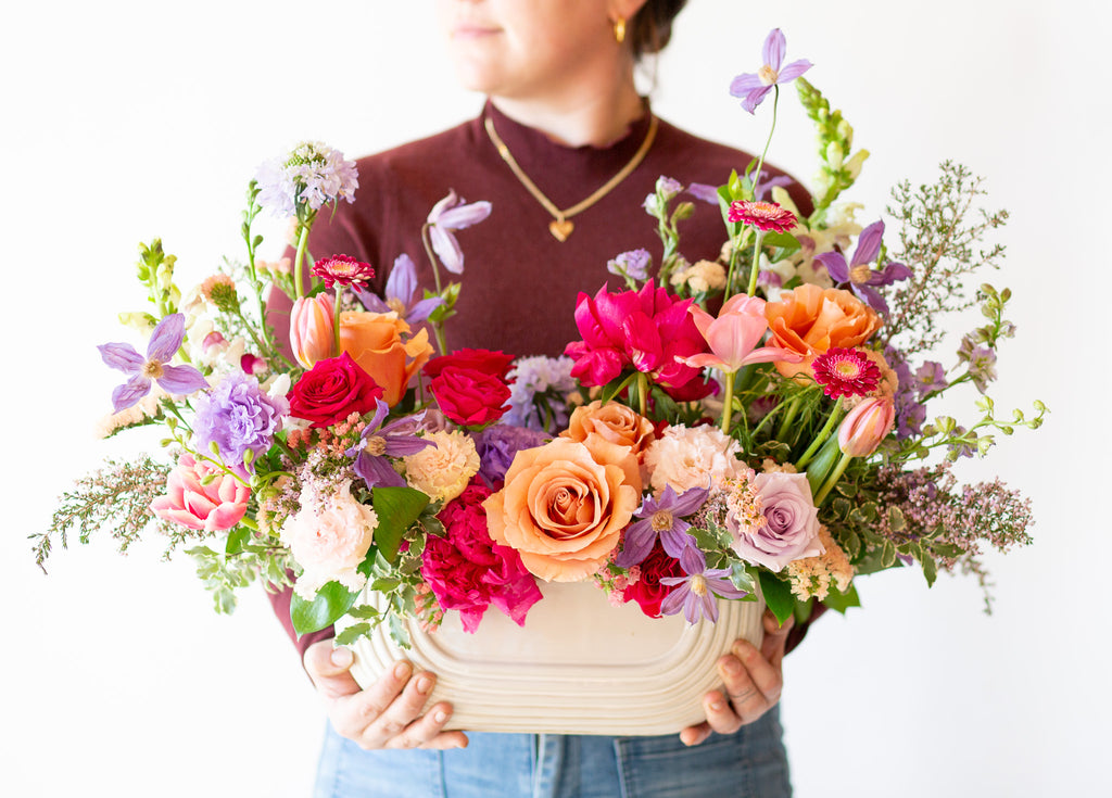 A woman holds a WildFlora's Wild Berry Garden Long & Low Arrangement in a white ceramic art deco long & low vase in front of a white backdrop. It has periwinkle, pink, magenta, orange, peach, purple and and white flowers, and includes rose, gerber daisy, lisianthus, snapdragon, clematis, scabiosa, peony, and tulips.