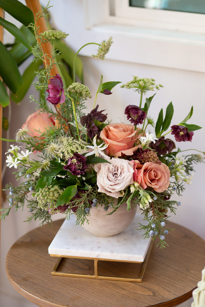 WildFlora's Winter's Garden Arrangement in a spherical grey vase, on a side table in a home. It has sienna orange, pastel violet, red, pink, fuchsia, eggplant purple, and white flowers, and includes rose, scabiosa, hellebore, Queen Anne's lace, and juniper.