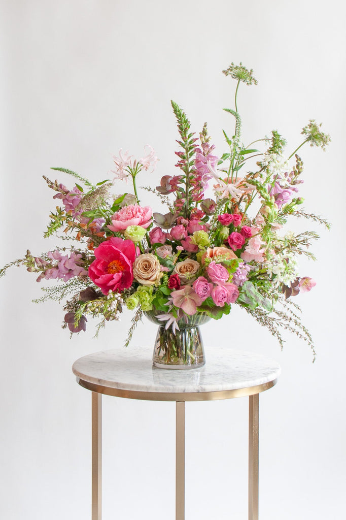 WildFlora's Secret Garden Flower Arrangement in a smokey glass compote vase on a small marble table in front of a white backdrop. It has dark purple, magenta, pink, chartreuse, orange, & white flowers, including peony, rose, spray rose, clematis, allium, hellebore, snapdragon, Queen Anne's lace, and lily.