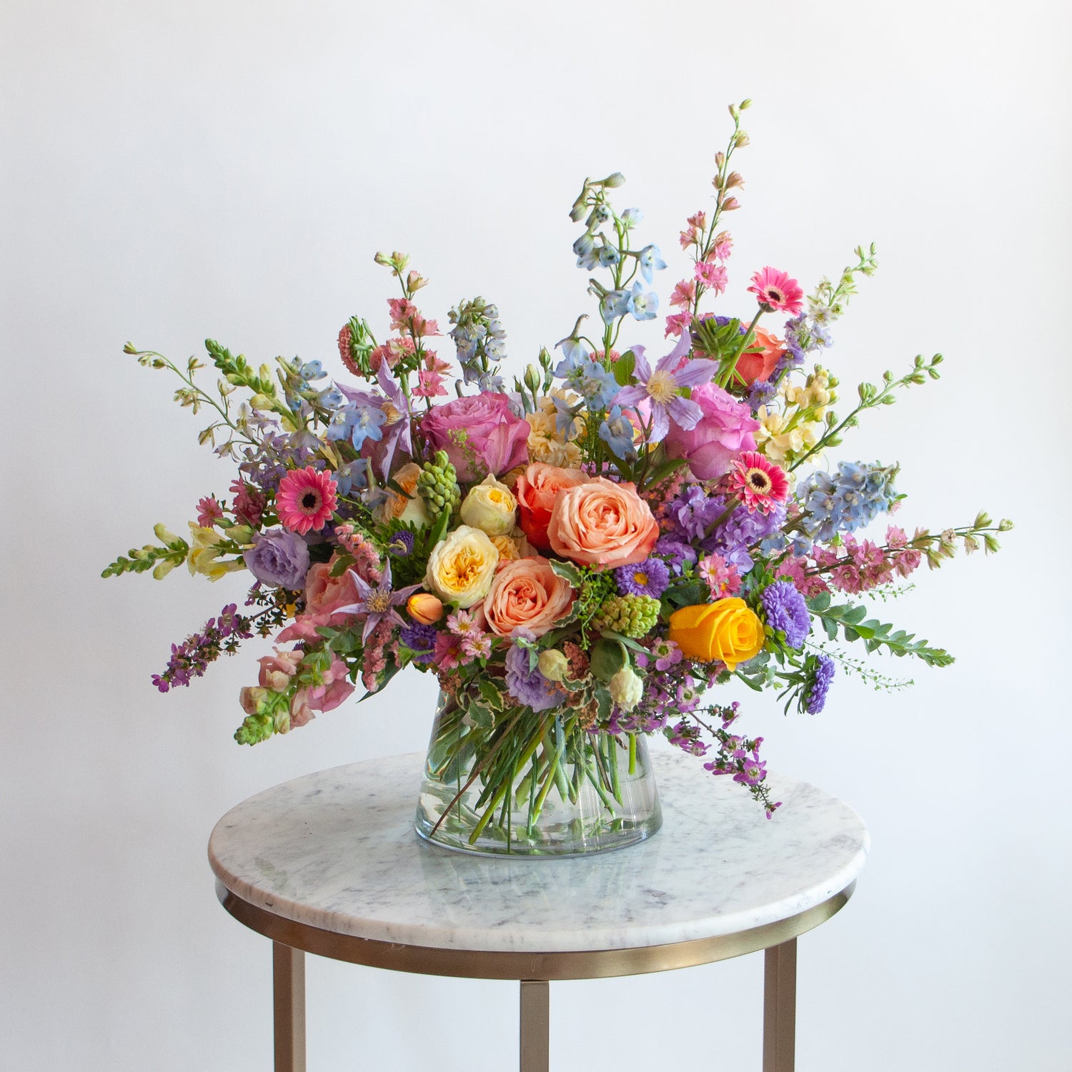A glass vase with a medium flower arrangement sits on a marble table in front of a white backdrop. The flowers are many colors and include roses, tulips, daisies, snapdragon, clematis, stock, hyacinth, and delphinium.