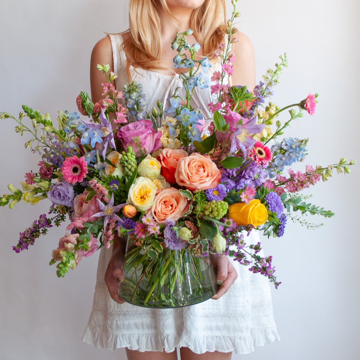 a woman in a white dress holds a glass vase with a large flower arrangement in it. The flowers are many colors and include roses, tulips, daisies, snapdragon, clematis, stock, hyacinth, and delphinium.