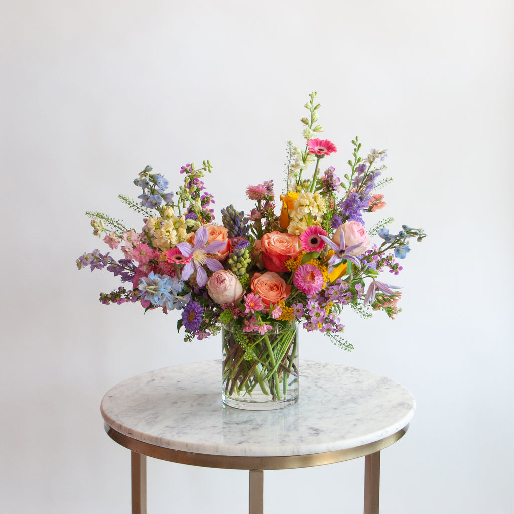 A glass vase with a medium flower arrangement sits on a marble table in front of a white backdrop. The flowers are many colors and include roses, tulips, daisies, snapdragon, clematis, stock, straw flower, and delphinium.