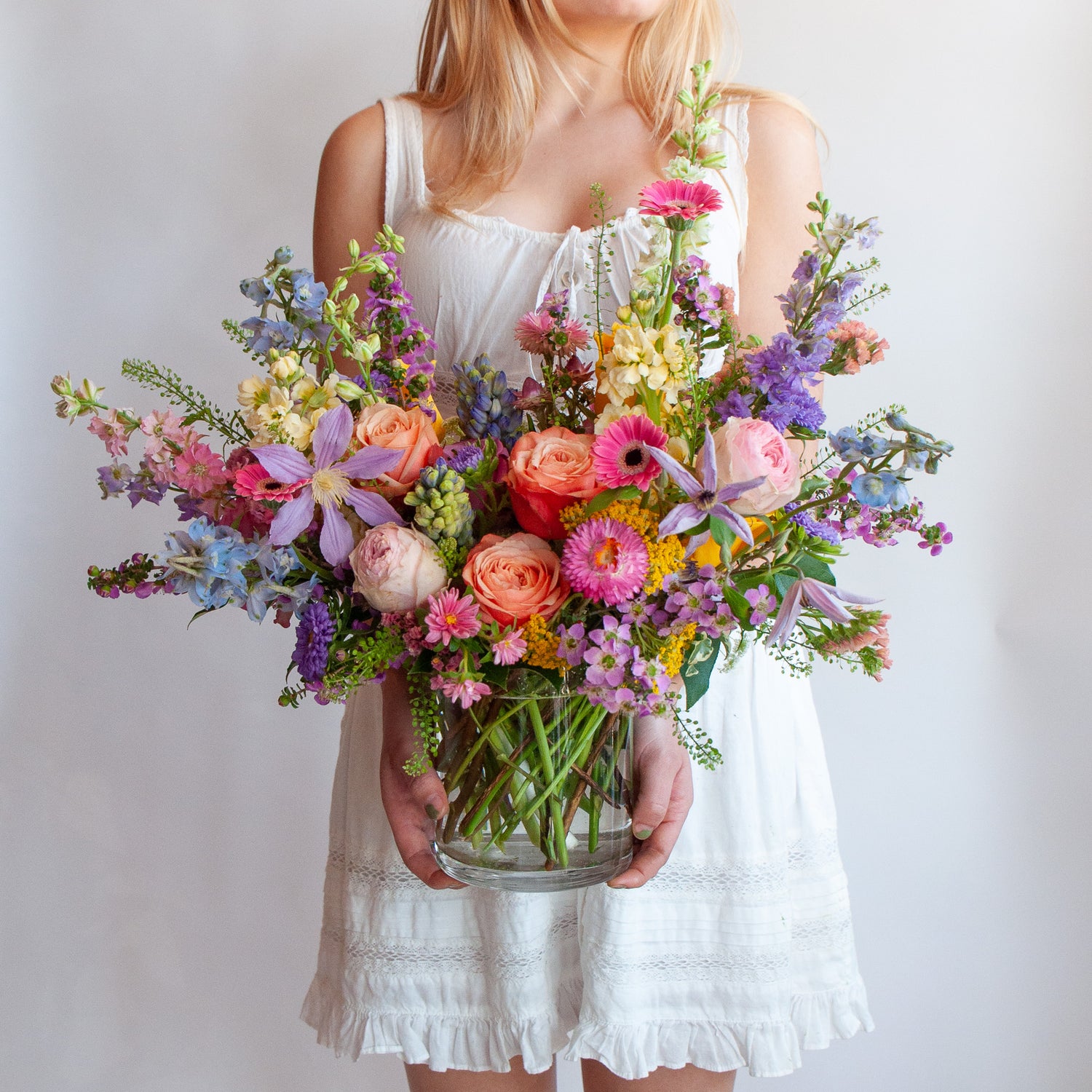 a woman in a white dress holds a glass vase with a medium flower arrangement in it. The flowers are many colors and include roses, tulips, daisies, snapdragon, clematis, stock, straw flower, and delphinium.