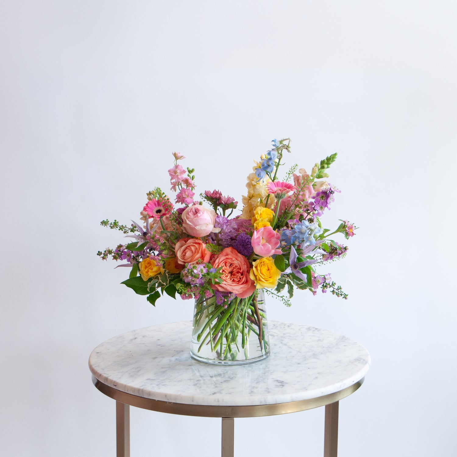 a glass vase with a small flower arrangement sits on a marble table in front of a white backdrop. The flowers are many colors and include roses, tulips, daisies, snapdragon, clematis, and delphinium.
