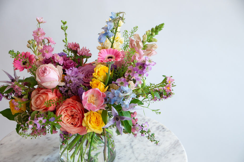 Close up of the blooms in a flower arrangement. The flowers are many colors and include roses, tulips, daisies, snapdragon, clematis, stock, and delphinium.