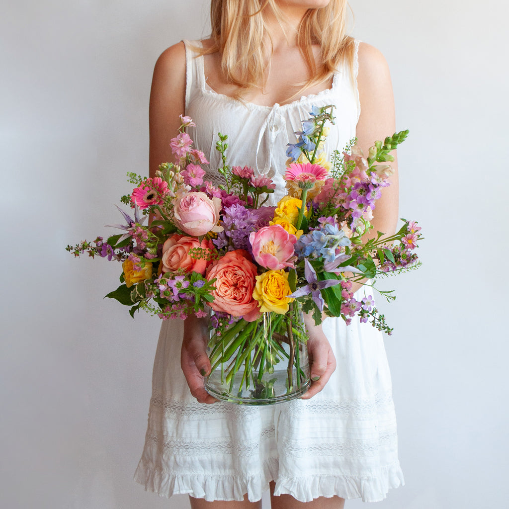 a woman in a white dress holds a glass vase with a small flower arrangement in it. The flowers are many colors and include roses, tulips, daisies, snapdragon, clematis, and delphinium.
