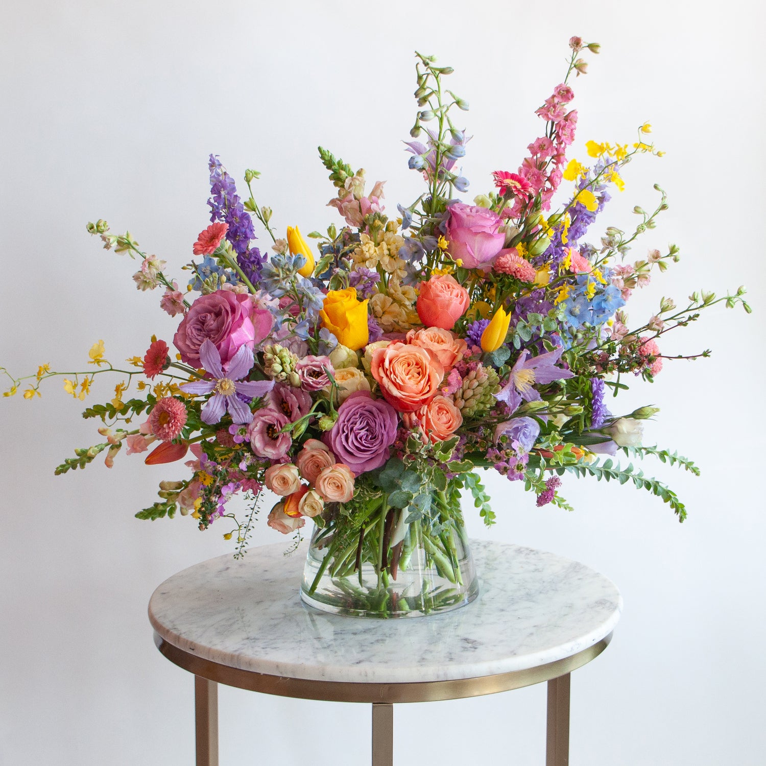 A glass vase with a medium flower arrangement sits on a marble table in front of a white backdrop. The flowers are many colors and include roses, tulips, daisies, snapdragon, clematis, stock, hyacinth, orchids, and delphinium.
