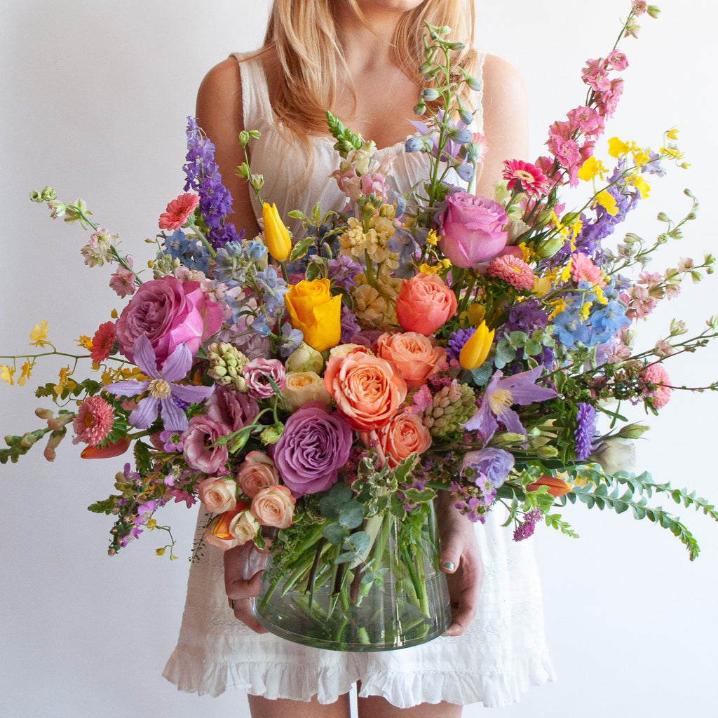a woman in a white dress holds a glass vase with an extra large flower arrangement in it. The flowers are many colors and include roses, tulips, daisies, snapdragon, clematis, stock, hyacinth, orchids, and delphinium.
