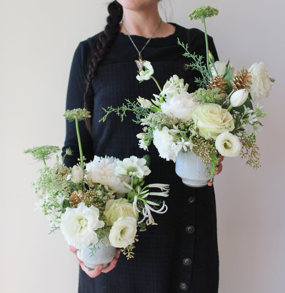 A woman holds WildFlora's Frost-Kissed Arrangement Pair in front of a white backdrop. They have green, white, and gold flowers, and include rose, Queen Anne's lace, pine cones, lily, scabiosa, tulip, and peony.