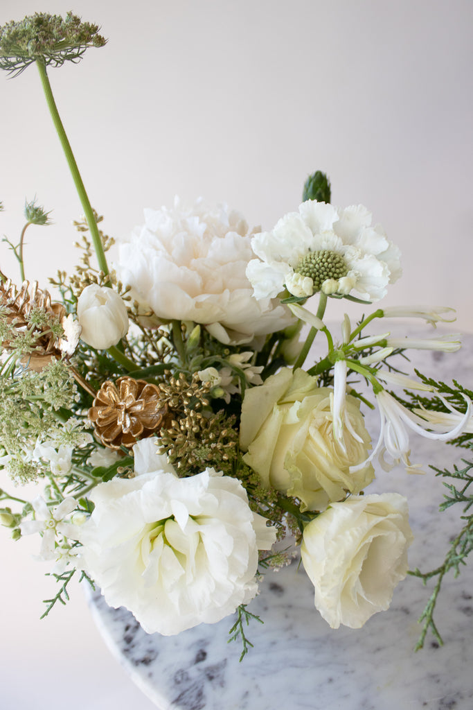 WildFlora's Frost-Kissed Arrangement Pair on a small marble table in front of a white backdrop. They have green, white, and gold flowers, and include rose, Queen Anne's lace, pine cones, lily, scabiosa, tulip, and peony.