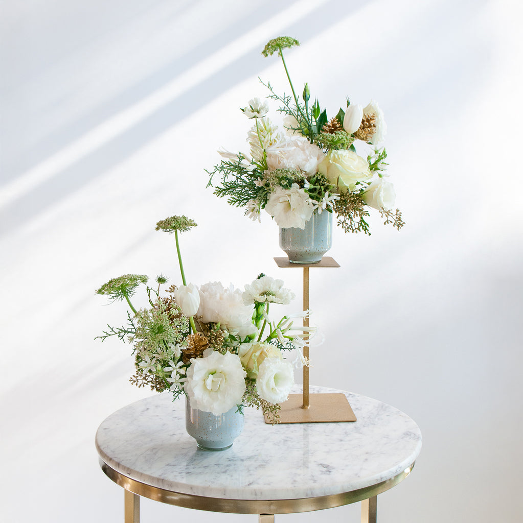 WildFlora's Frost-Kissed Arrangement Pair on a small marble table in front of a white backdrop. They have green, white, and gold flowers, and include rose, Queen Anne's lace, pine cones, lily, scabiosa, tulip, and peony.