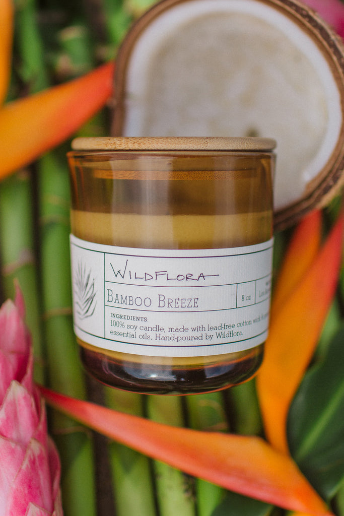 Bamboo Breeze candle in a golden-tan glass jar with a wood lid on a bed of bamboo with a coconut, and ginger and heliconia flowers