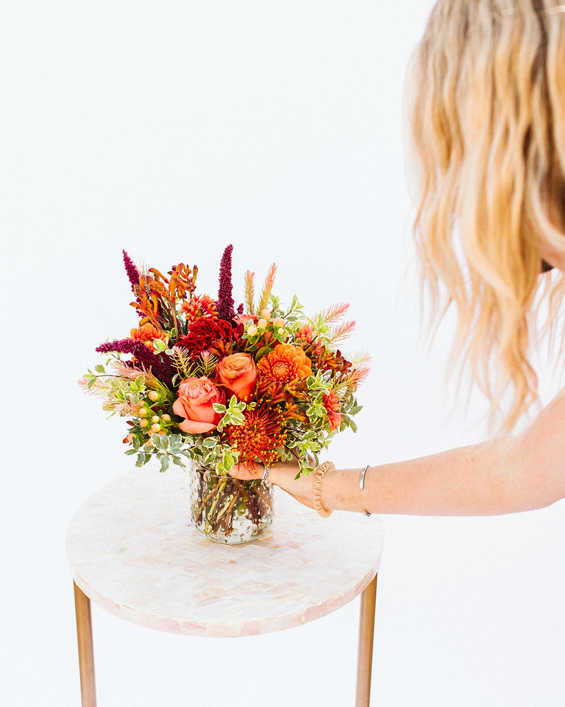 A hand holding a vibrant autumn floral arrangement in a clear glass vase, set on a round light pink marble side table against a solid white background.