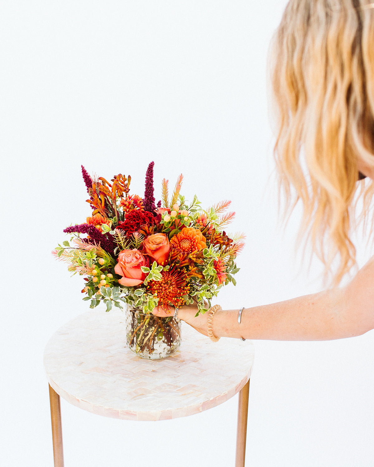 A hand holding a vibrant autumn floral arrangement in a clear glass vase, set on a round light pink marble side table against a solid white background.