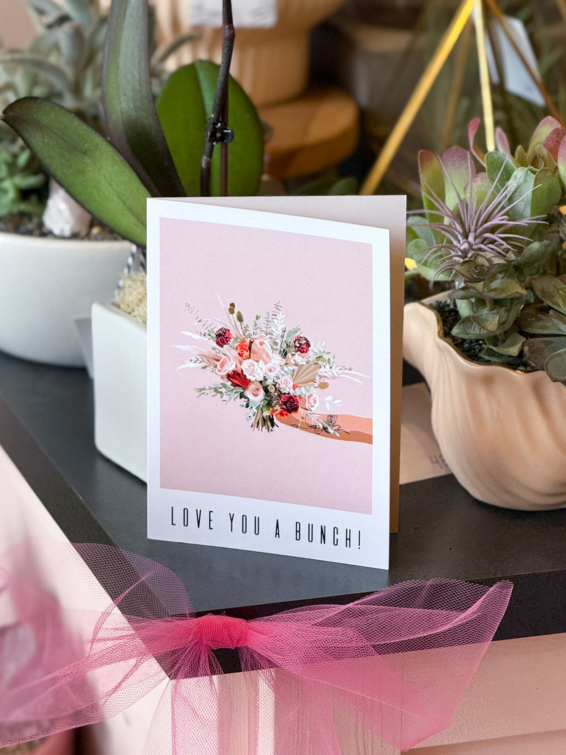 Greeting card with floral design and 'Love You a Bunch!' text, placed on a surface with plants.