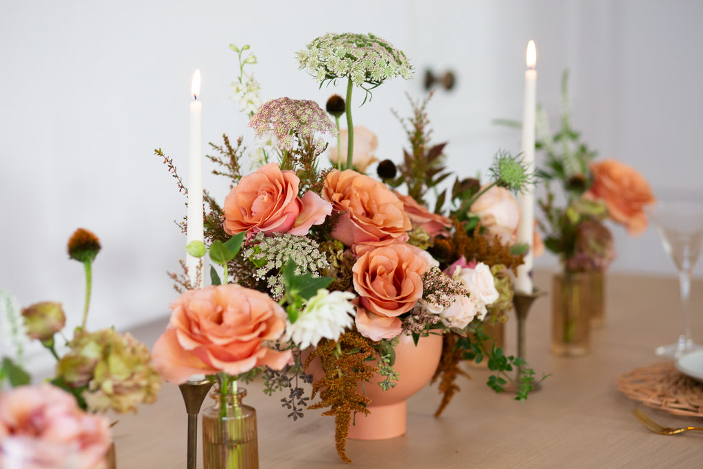 WildFlora's Gathered Garden Arrangement in a terracotta compote container & Bud Vases in tan ribbed glass vases a on Thanksgiving table. It includes dusty coral-orange, blush, brown, and tan earth-toned flowers, complete with Moab roses, spray roses, weeping amaranthus, Queen Anne's lace, lisianthus, echinacea, and foliage. 