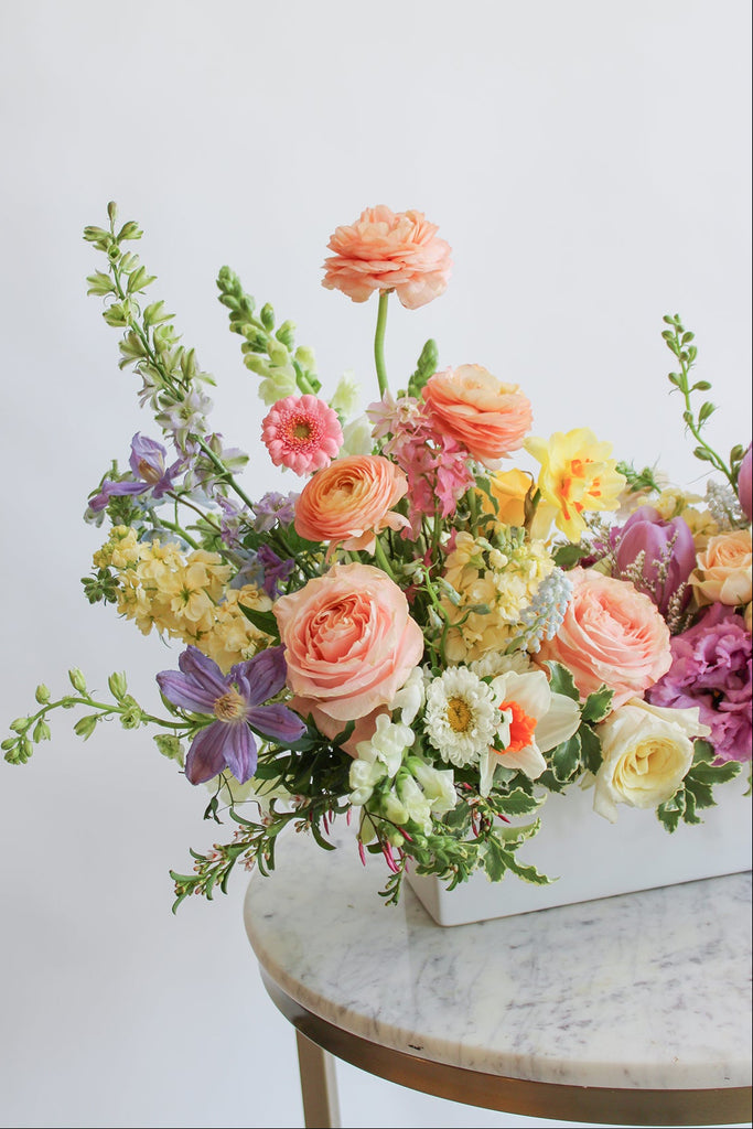 A long and low floral arrangement held by a person, featuring a variety of flowers including purple peach yellow and white blooms and green foliage.