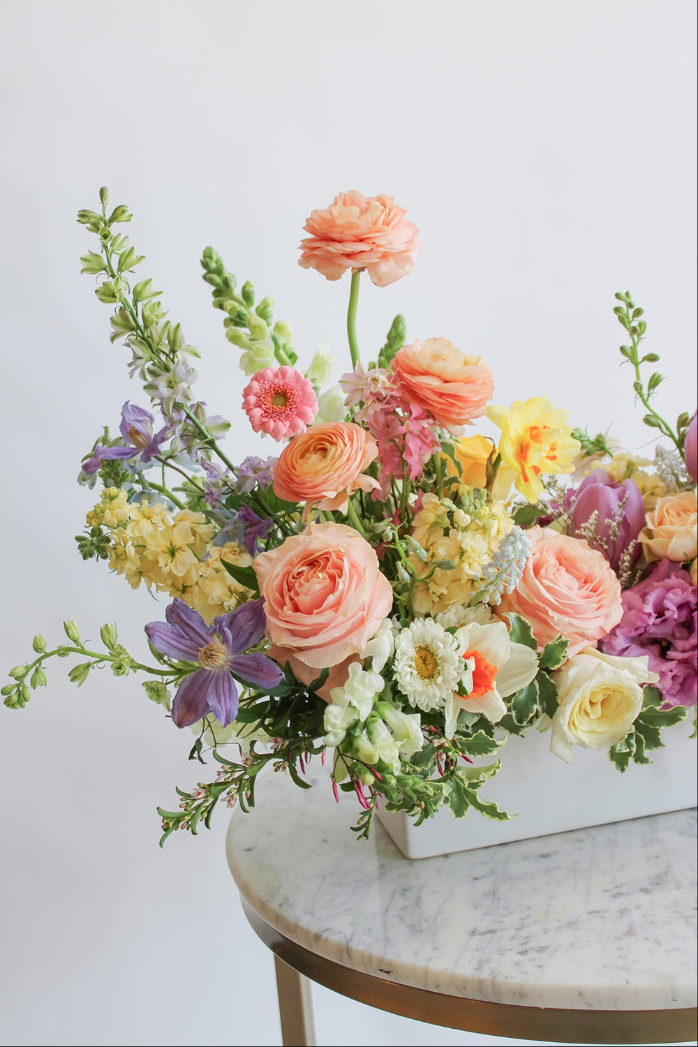 A long and low floral arrangement held by a person, featuring a variety of flowers including purple peach yellow and white blooms and green foliage.