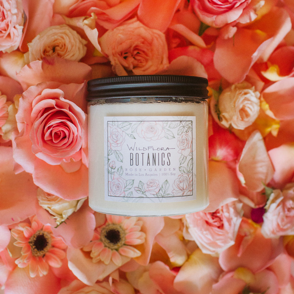 Rose Garden Botanics double-wick, white soy wax candle in a glass container with a black lid. The label has a matching botanical sketch and shows the scent. Behind the candle are pink roses