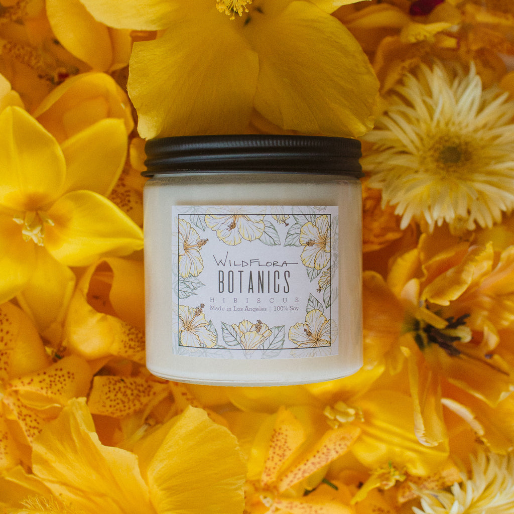 Hibiscus Botanics double-wick, white soy wax candle in a glass container with a black lid. The label has a matching botanical sketch and shows the scent. Behind the candle are yellow flowers