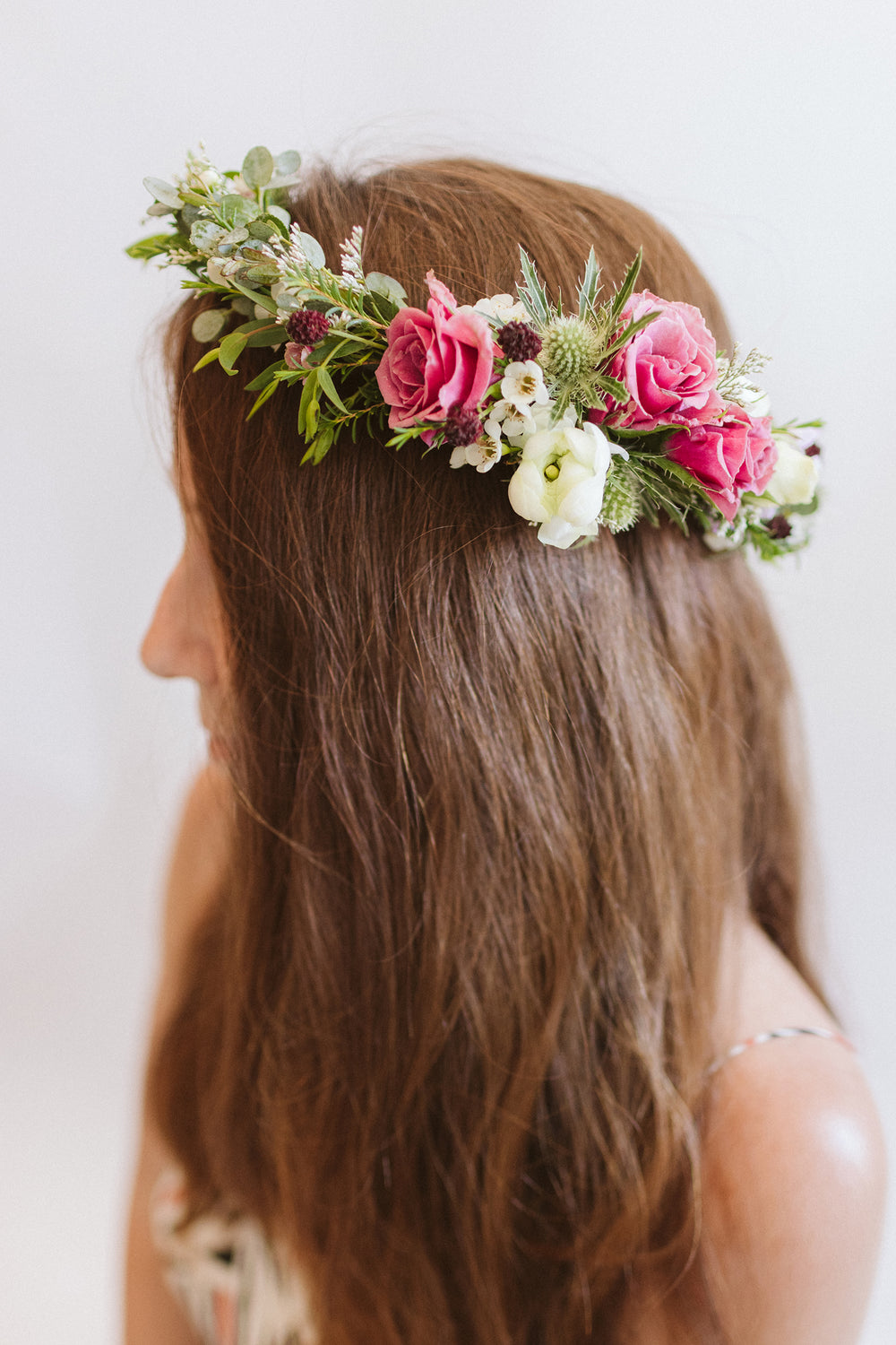 a profile of woman with reddish brown hair wears a flower crown in front of a white backdrop. It's made of pink, white, green, and dark purple flowers, including spray rose, thistle, ranunculus, and other foliage.