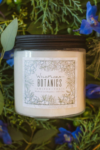 Juniper Trail Botanics double-wick, white soy wax candle in a glass container with a black lid. The label has a matching botanical sketch and shows the scent. Behind the Candle are evergreens and blue flowers