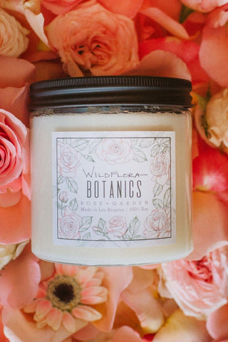 Rose Garden Botanics double-wick, white soy wax candle in a glass container with a black lid. The label has a matching botanical sketch and shows the scent. Behind the candle are pink roses